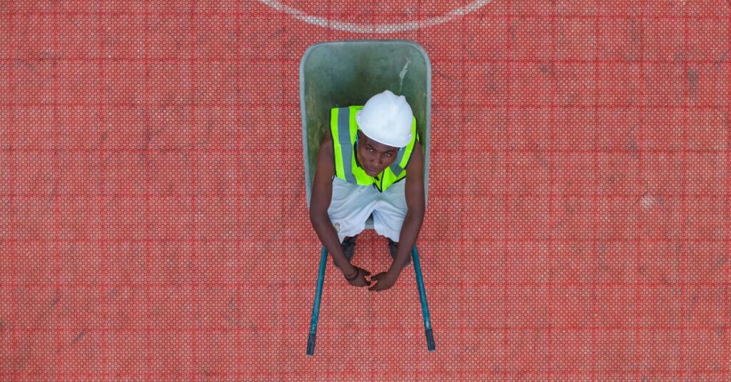 A construction worker in safety gear sits in a wheelbarrow on a textured red surface.
