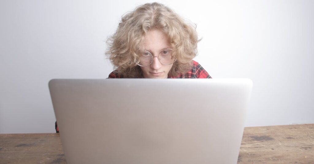 Young adult with curly hair working intently on a laptop in a minimalist indoor setting.