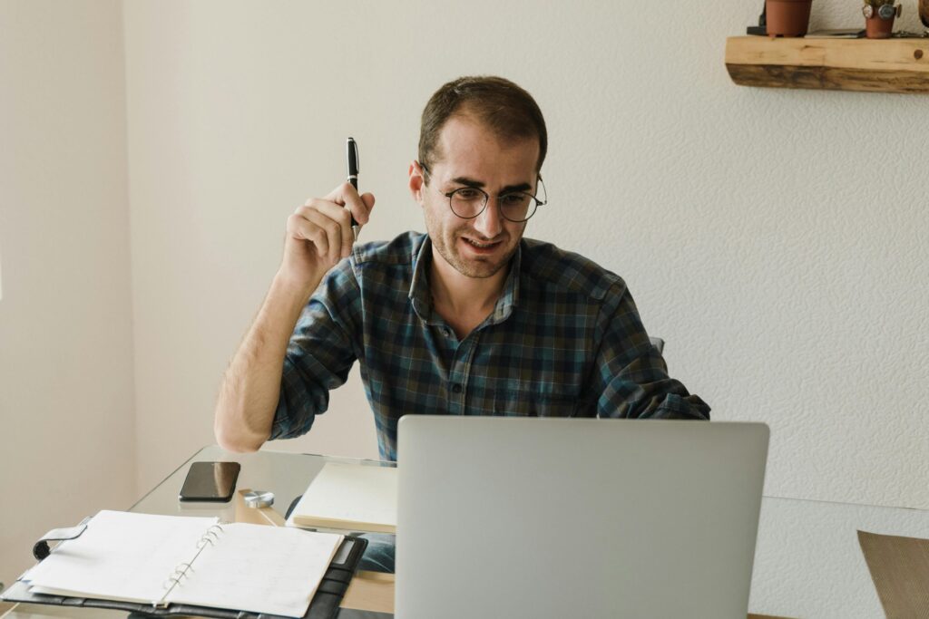 A man working remotely at home using a laptop, surrounded by notebooks and a smartphone.