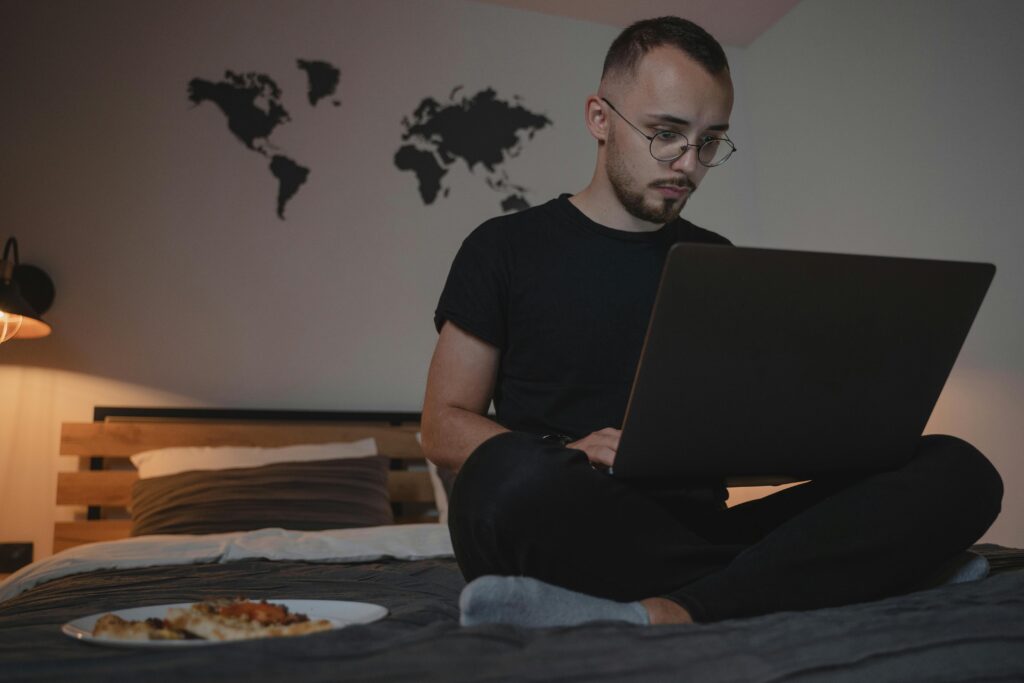 A man sits on a bed with a laptop, working remotely, with a world map and pizza plate nearby.