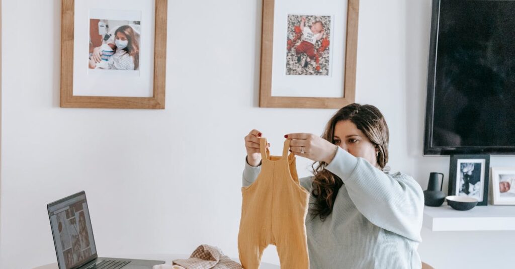 Happy young mother with long hair in casual sweatshirt folding clothes of baby while sitting at table with laptop in modern light apartment