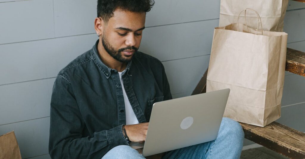 Young man using a laptop surrounded by shopping bags and packages, working on eCommerce business.