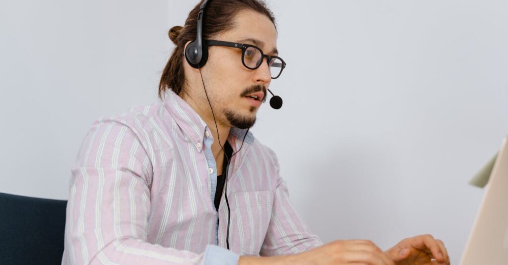 A young man in a striped shirt works at a laptop, wearing a headset for customer support.