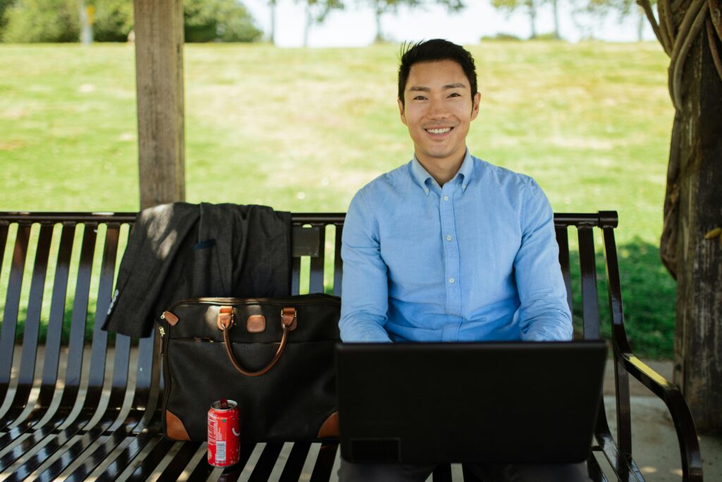 Asian man in casual attire working on laptop outside on a sunny day, enjoying remote work lifestyle.