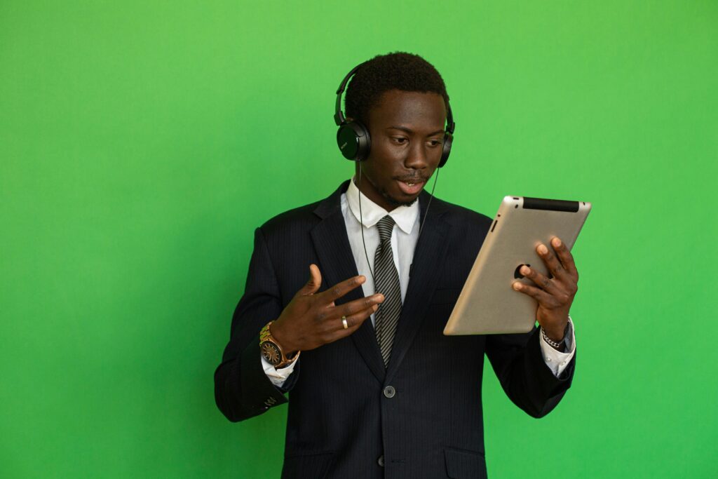 Young black man in formal attire using a tablet with headphones against a green background.