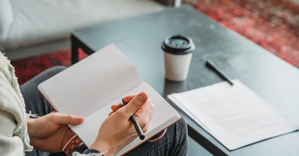 Close-up of a person writing in a notebook, with coffee on a table, indoors.