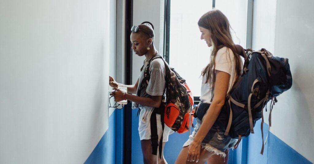 Two women travelers in a hostel corridor with backpacks, preparing for their journey.